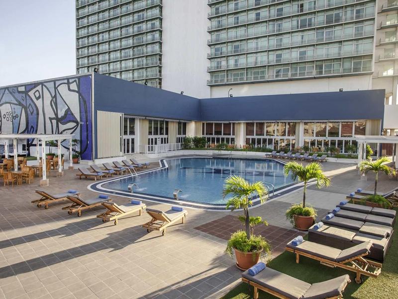 Moderner Außenpool mit Liegestühlen, Pflanzen und hohen Gebäuden im Hintergrund bei klarem Himmel.