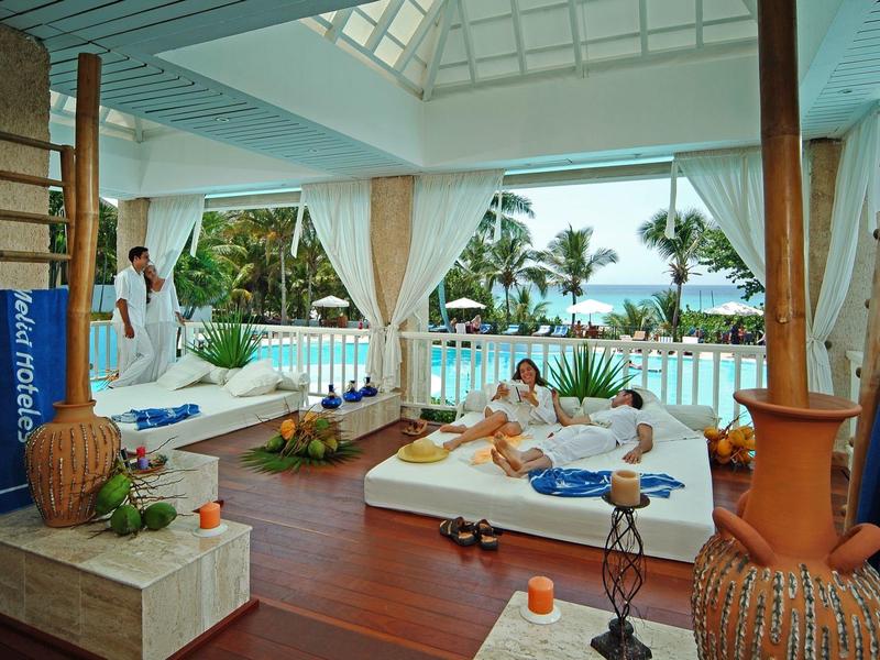 Moderner Wellnessbereich mit Liegen, Holzterrasse, großen Vasen und Pool vor tropischer Pflanzenkulisse.