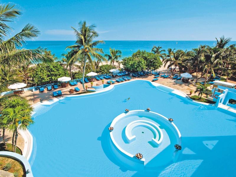 Großer blauer Pool mit runden Inseln, Palmen, Sonnenschirmen und Blick aufs Meer im Hintergrund.