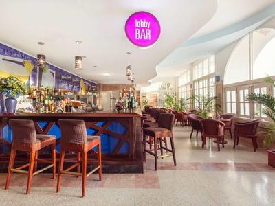 Helle Bar mit Holzmöbeln, großen Fenstern, Pflanzen und gemütlichen Sitzbereichen.