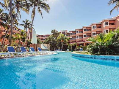 Großer Außenpool umgeben von Liegestühlen und Palmen vor einem Hotelgebäude.