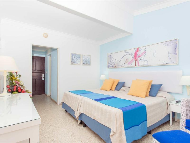 Hell eingerichtetes Schlafzimmer mit zwei Betten, blauen und gelben Akzenten und weißem Mobiliar.