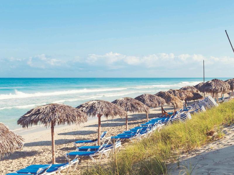 Strand mit Strohschirmen und blauen Liegestühlen am Meer unter klarem Himmel.