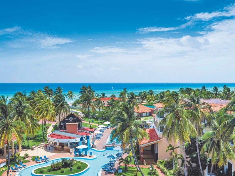 Blick auf tropische Hotelanlage mit Pool, Palmen und Meer unter blauem Himmel.