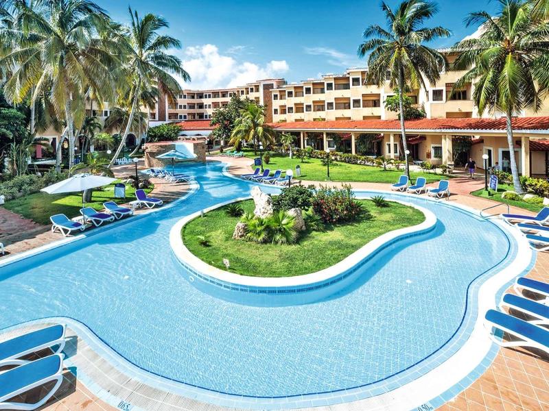 Großer Hotelpool mit tropischer Insel und Liegestühlen unter Palmen bei sonnigem Wetter.