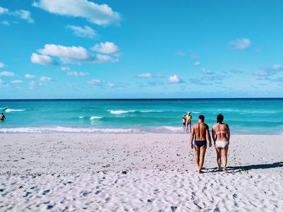 Zwei Personen gehen am sonnigen Strand zum Meer mit blauem Himmel und vereinzelten Wolken.