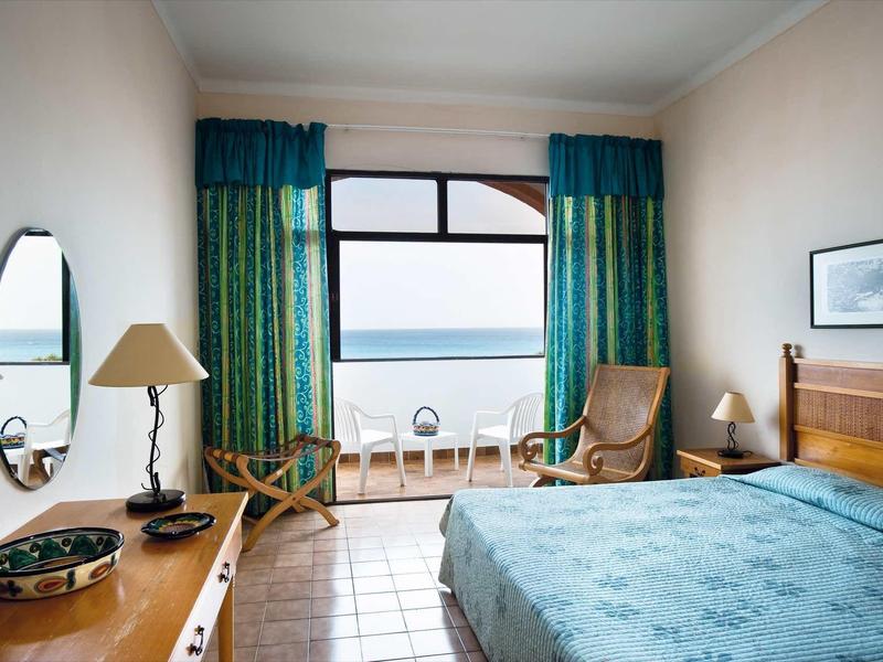 Camera d'hotel luminosa con letto matrimoniale, scrivania, due poltrone e balcone con vista mare.