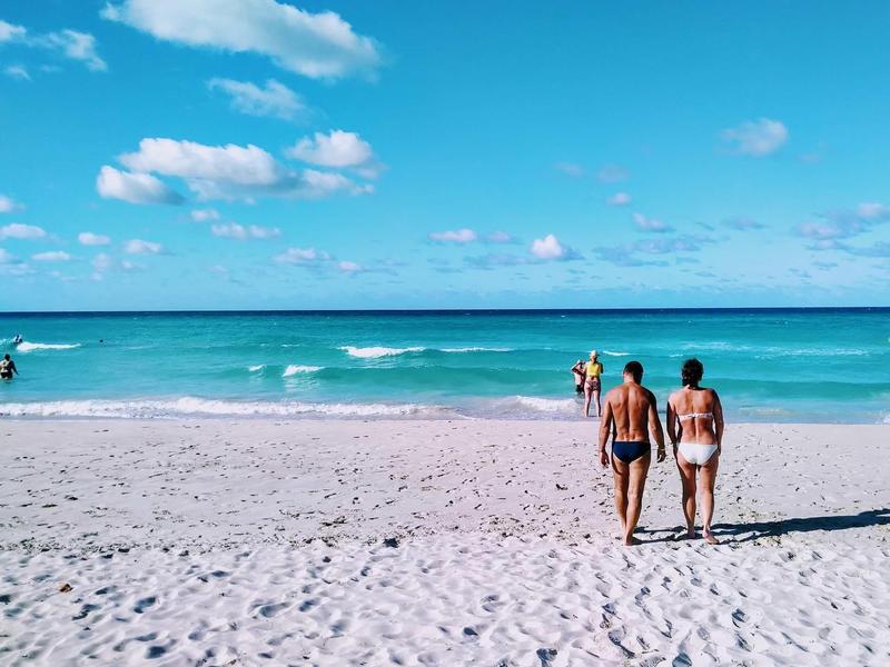 Due persone camminano su una spiaggia di sabbia bianca con cielo e mare blu.