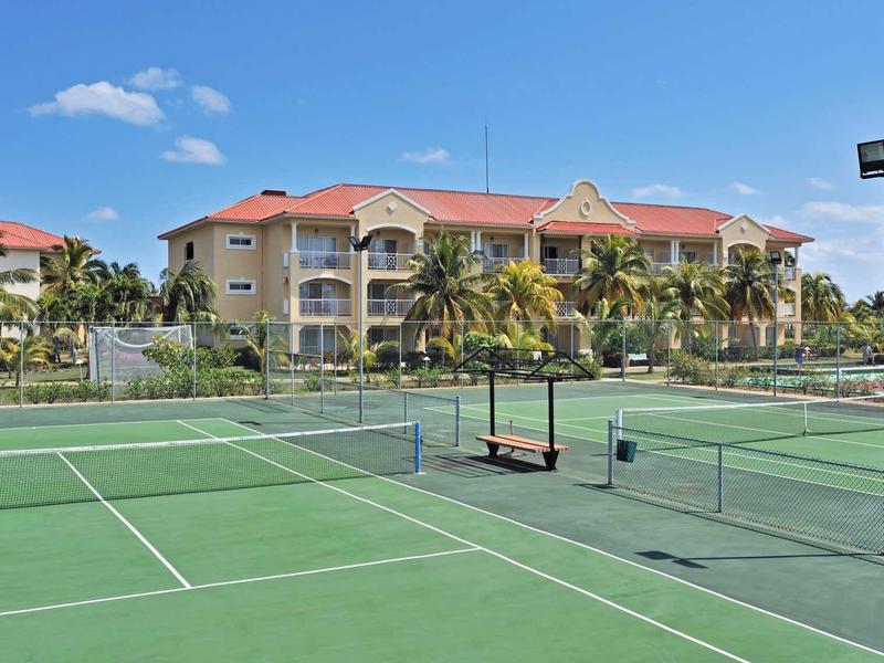 Campo da tennis verde di fronte a un edificio alberghiero con palme sotto un cielo azzurro.