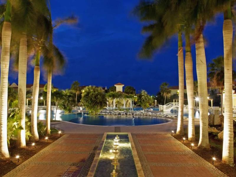 Viale illuminato che conduce a una piscina circondata da palme di notte.