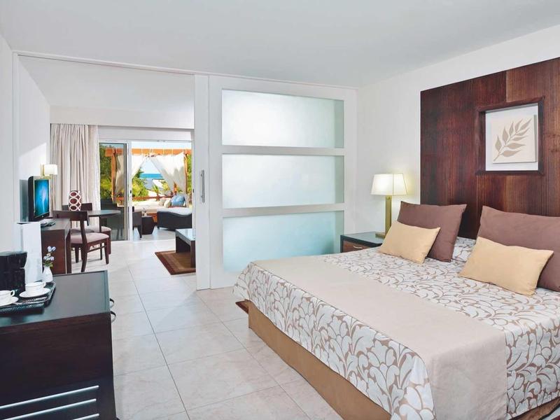 Camera d'hotel moderna con letto grande, TV, comodini e accesso a un soggiorno luminoso.