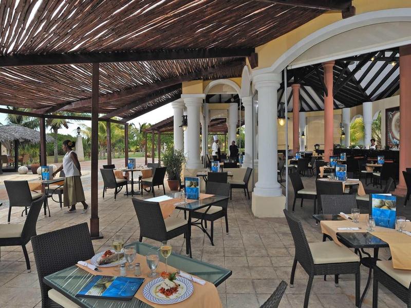 Terrazza del ristorante con tavoli quadrati, sedie in vimini e tetto in legno sparso.