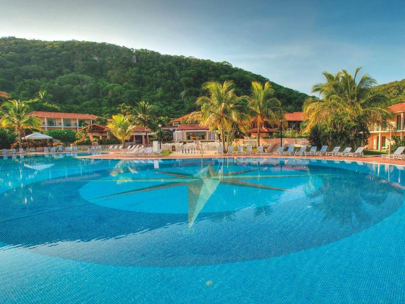 Großer Pool mit Sonnenschirmen und Hotels im Hintergrund, umgeben von grünen Hügeln.