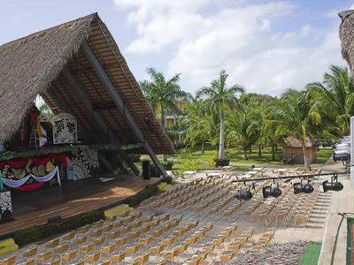 Freiluftbühne mit einem überdachten Bereich, Sitzreihen und tropischer Vegetation im Hintergrund.