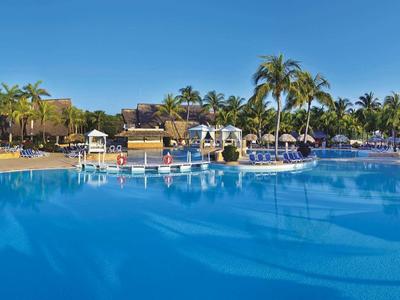 Großer blauer Pool mit Palmen und Liegestühlen unter klarem Himmel in einer tropischen Umgebung.