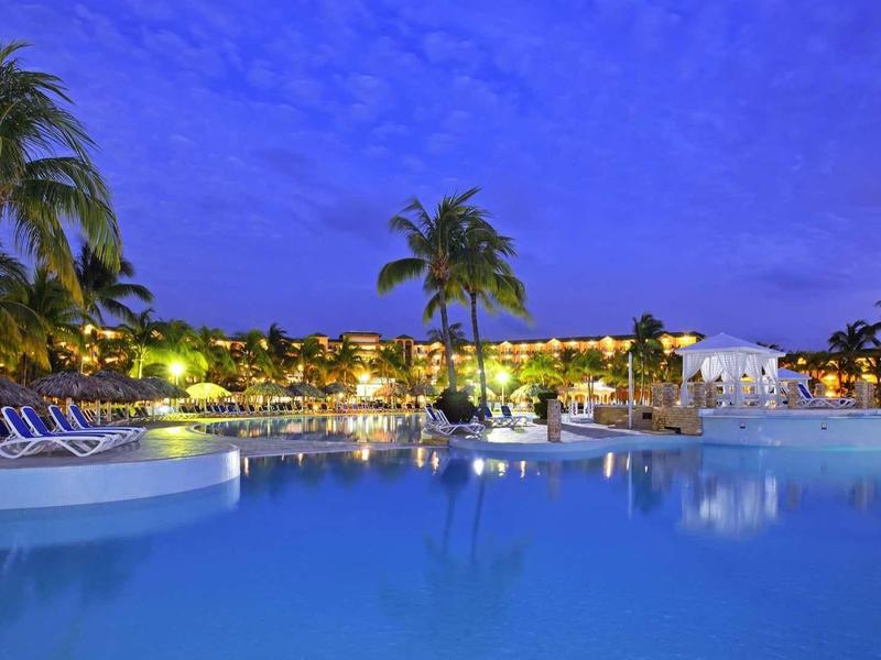 Noční osvětlený hotelový bazén s palmami a lehátky.