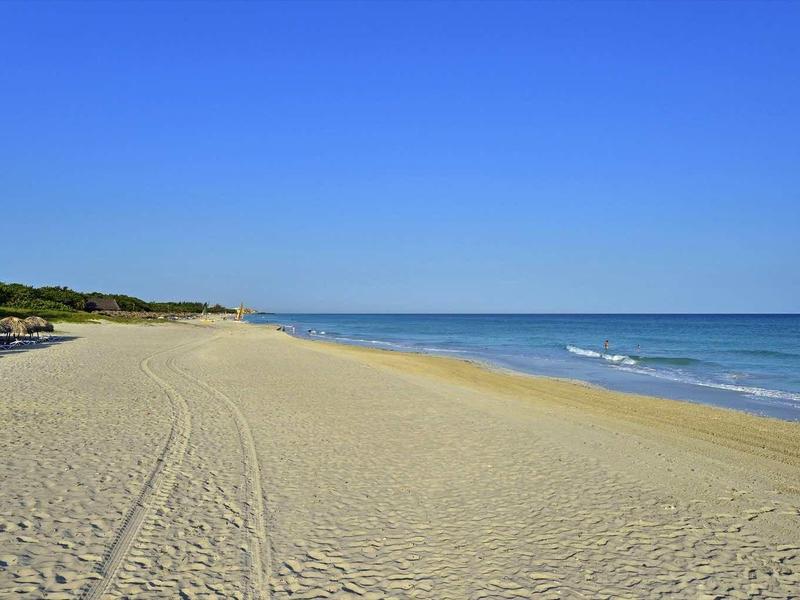 Pláž s jemným pískem, slunečníky a modrým mořem pod jasnou oblohou.