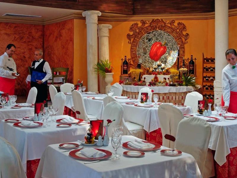 Elegantní místnost s bílými stoly, červenými ubrusy, číšníky v uniformách a dekorativním zrcadlem na stěně.