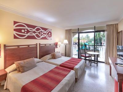 Hotelový pokoj se dvěma jednolůžky, moderním nábytkem a balkonem s výhledem na palmy.