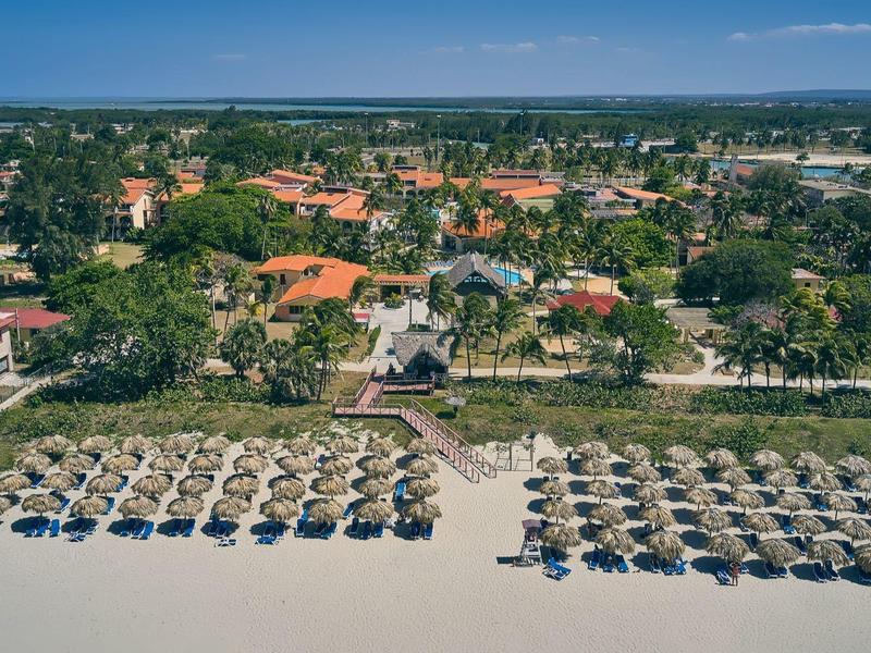 Veduta aerea di una spiaggia con ombrelloni, lettini e un complesso alberghiero sullo sfondo.
