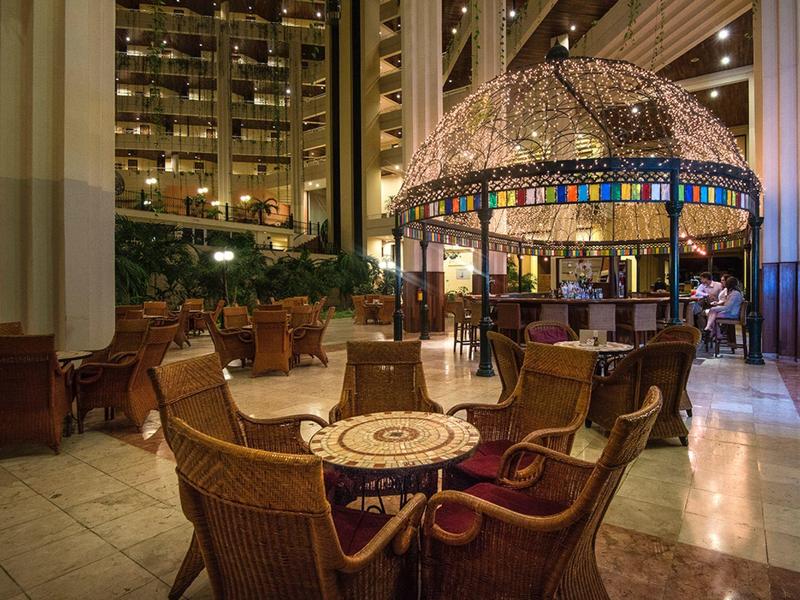 Lounge dell'hotel con tavoli rotondi, sedie in vimini e un bar con cupola di vetro colorato sullo sfondo.