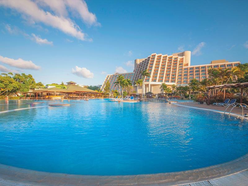 Grande piscina blu davanti a un hotel sotto un cielo azzurro con poche nuvole.