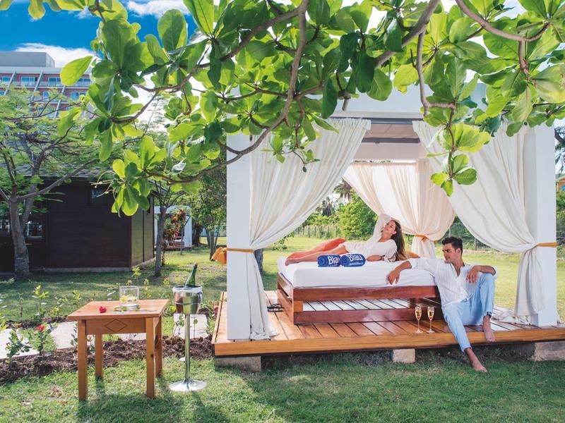 Un letto all'aperto con tende bianche su una piattaforma di legno in un giardino verde con due persone.