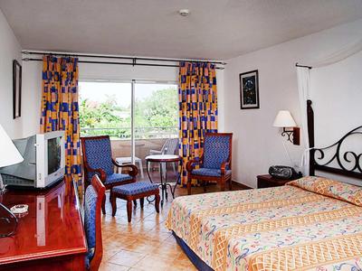 Hotel room with double bed, two armchairs, TV, and window view of green trees.