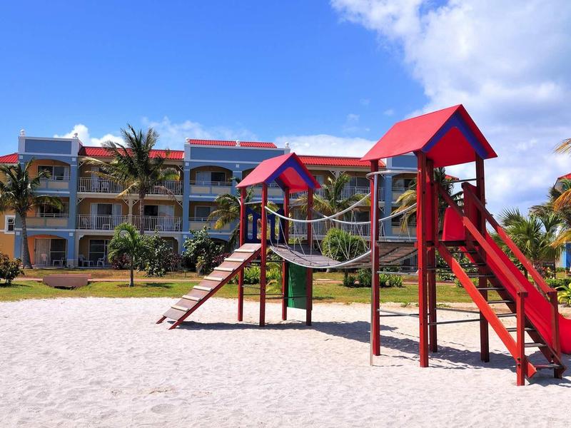 Playground with climbing frame and slide in front of a hotel building under a bright sky.