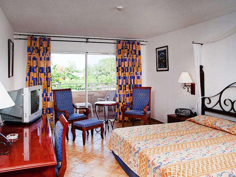 Hotel room with double bed, two armchairs, TV, and window view of green trees.