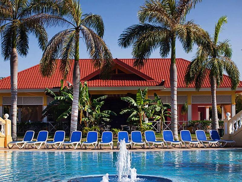 Hotel with red roof, palm trees, and pool with blue lounge chairs in sunny weather.