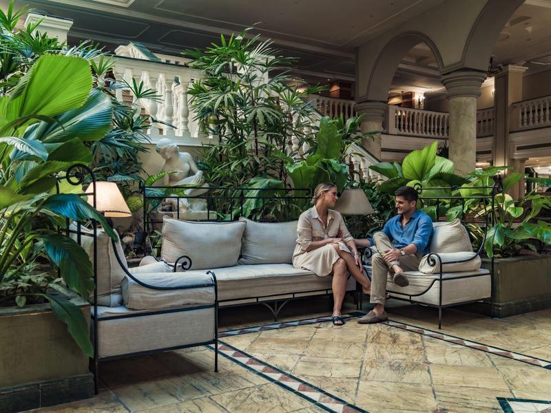 Twee mensen zitten op een bank in een hotelentree vol planten met lichte vloer.