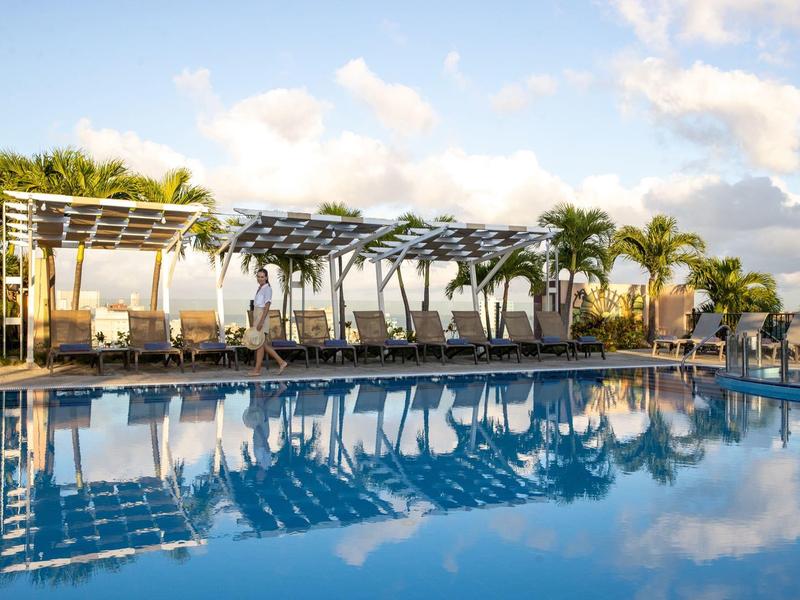 Groot zwembad met zonneschermen en palmbomen onder een blauwe hemel bij een resort.