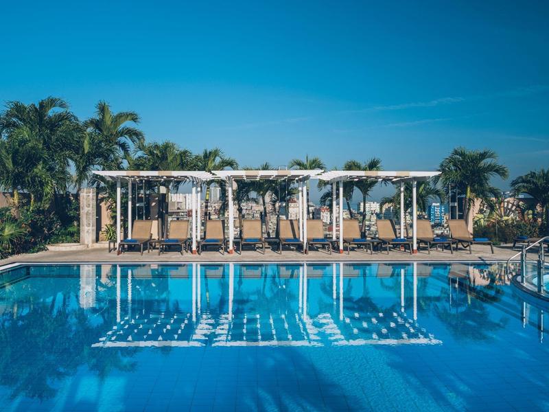 Groot zwembad met ligstoelen en palmbomen onder een heldere lucht bij een hotelresort.