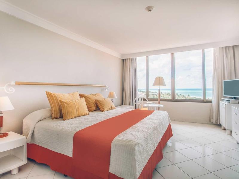 Camera d'hotel luminosa con letto grande, copriletto arancione e vista sul mare dalla grande finestra.