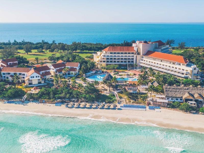 Struttura alberghiera sulla spiaggia con ombrelloni e vista sul mare in giornata limpida.