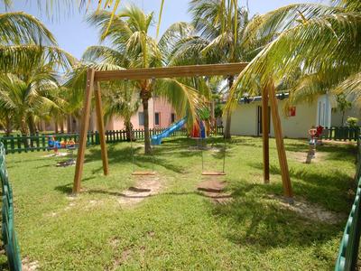 Ein Holzschaukelgestell mit zwei Sitzen steht auf grünem Rasen, umgeben von Palmen und kleinen Gebäuden.