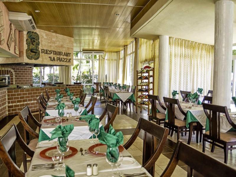 Restaurant mit Holzmöbeln, weißen Tischdecken und grünen Servietten in hellem Raum mit großen Fenstern.