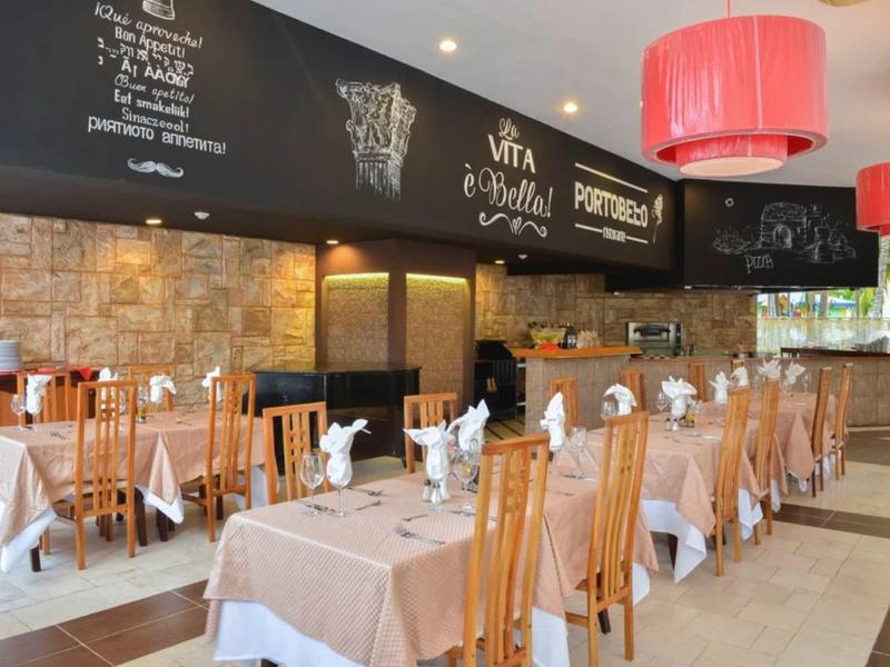 Helles Restaurant mit Holzstühlen, braunen Tischdecken, roten Lampenschirmen und Menütafel an der Wand.