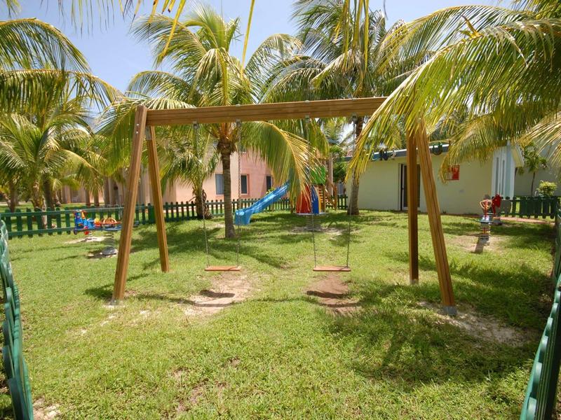 Ein Holzschaukelgestell mit zwei Sitzen steht auf grünem Rasen, umgeben von Palmen und kleinen Gebäuden.