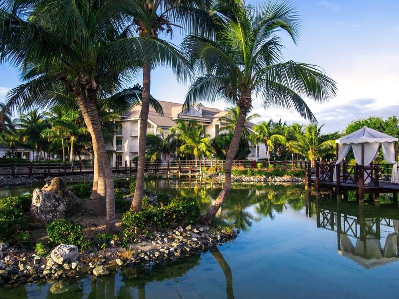 Complesso turistico tropicale con stagno, palme e gazebo sull'acqua calma.