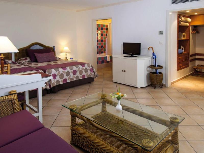 Camera d'hotel con letto matrimoniale, divano, tavolo di vetro, TV e arredamento accogliente.