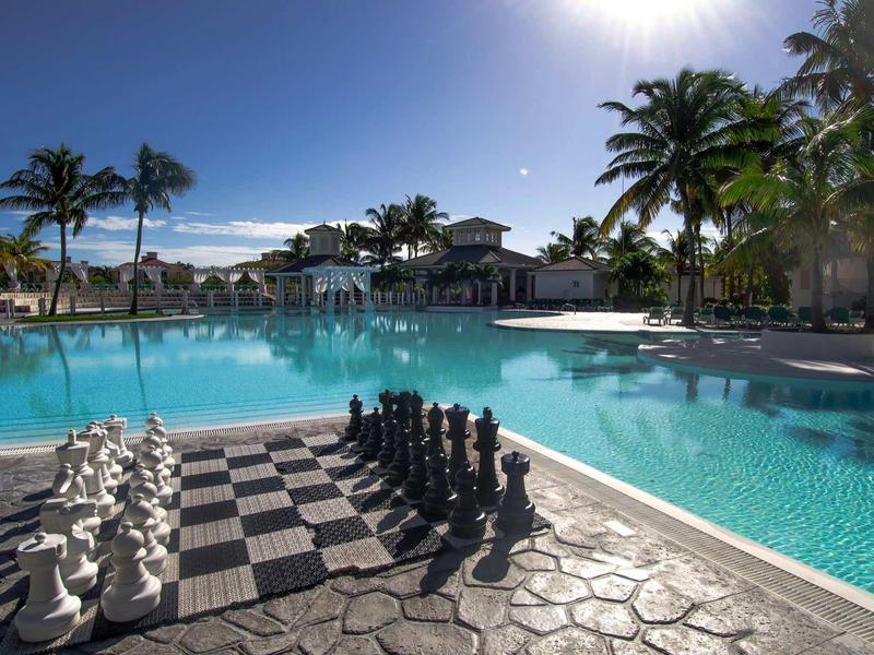 Grande scacchiera accanto a una piscina lussuosa con palme sotto un cielo limpido.