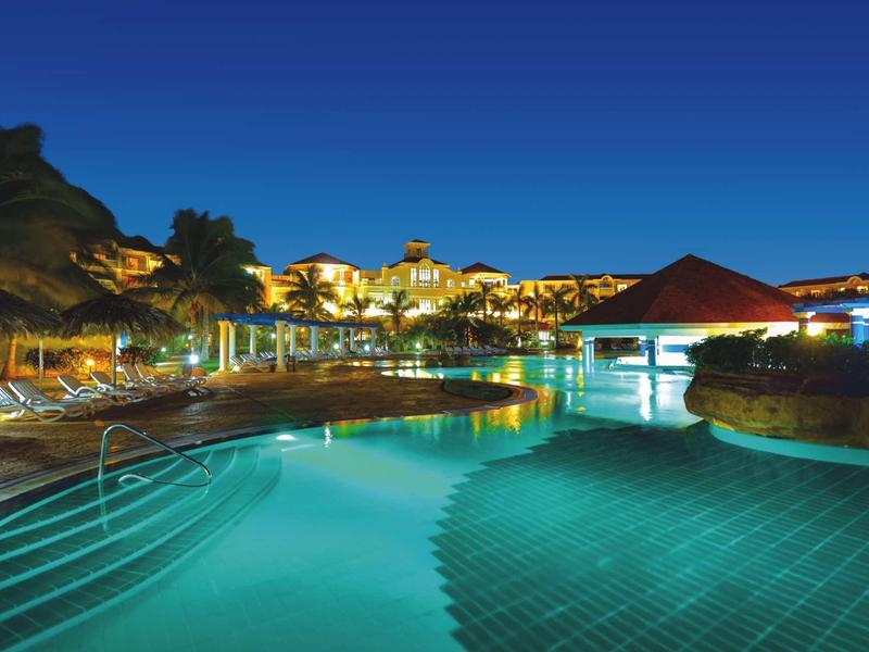 Grande piscina dell'hotel illuminata di notte con edifici circostanti e cielo limpido.