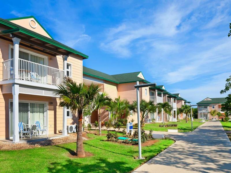 Rijen vakantieappartementen met balkons, omgeven door palmbomen en een pad onder een blauwe lucht.