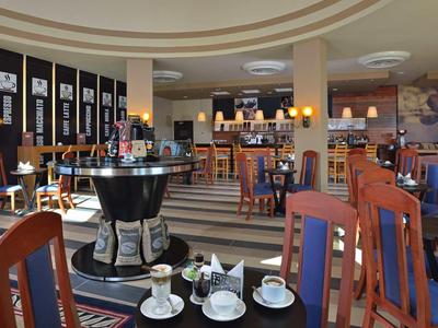 Jasně osvětlený hotelový salon s stolky, židlemi a kávovarem uprostřed.