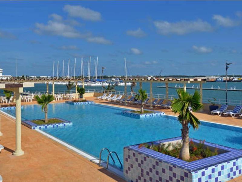 Grande piscina all'aperto con lettini e vista sul porto turistico in una giornata di sole.