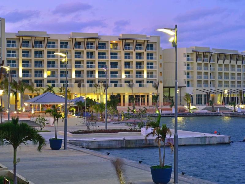 Complesso alberghiero con vista mare, giardino di palme e percorso illuminato sull'acqua al crepuscolo.