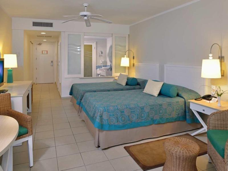 Camera d'hotel con due letti matrimoniali, area salotto, TV e scrivania in tonalità neutre.