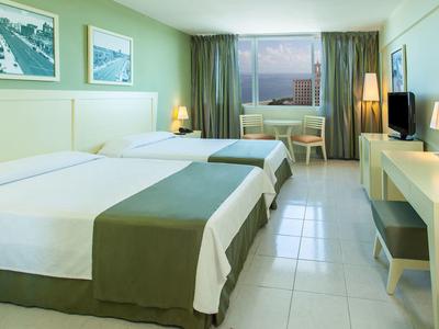 Chambre d'hôtel lumineuse avec deux lits simples, bureau, télévision et fenêtre avec vue extérieure.